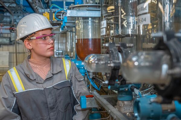 junge Frau mit Helm steht vor einer industriellen Anlage mit Zylindern