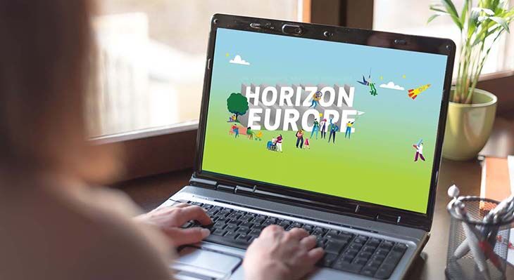 Das Bild zeigt verschwommen eine Person von hinten die ihre Hände auf einem Laptop hat, der offen steht. Auf dem Desktop ist das das Logo von "Horizon Europe" auf einem grünen Hintergrund voller animierter Menschen zu sehen.