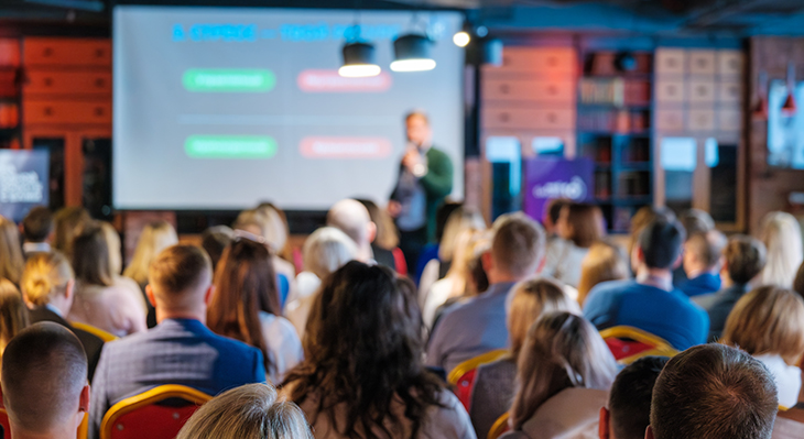 Konferenz mit einem Sprecher vor einer Leinwand. Das Publikum ist dem Sprecher zugewandt.