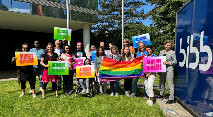 Auf dem Bild sind mehrere Personen zu sehen, die eine Regenbogenflagge und mehrere Plakate in der Hand halten. Auf den Plakaten steht das Wort Stimme.