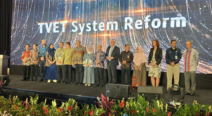 Prof Dr Esser stands on a black stage with a delegation. In the background you can read "TVET System Reform"