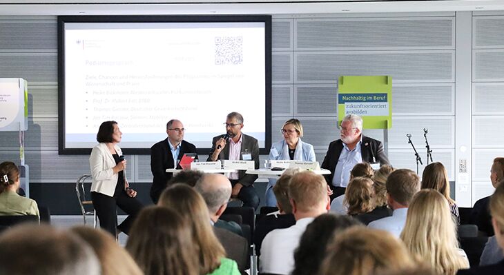 Moderiert Podiumsdiskussion mit 5 Teilnehmern vor einer großen Gruppe an Menschen.