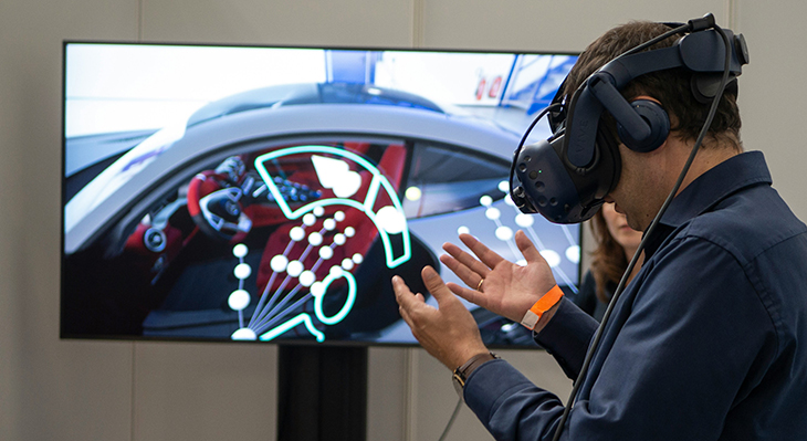 Mann mit blauer Jacke und VR-Brille steht vor einem Bildschirm, auf dem der innere Steuerbereich eines Autos zu sehen ist.