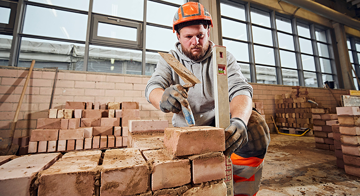 Ein Mann in Arbeitskleidung beim mauern einer Backsteinmauer: Er schaut konzentriert auf die Wasserwaage, im Vordergrund die ersten Reihen einer Mauer, hinter der er kniet. 