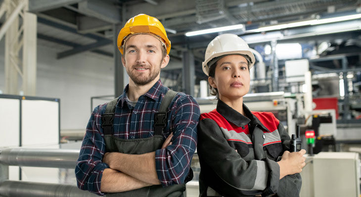 Zwei junge, Arbeiter mit Helmen und Arbeitskleidung stehen in einer großen Werkshalle