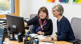 Zwei Frauen sitzen im Büro am Schreibtisch und betrachten einen Bildschirm. Die linke Frau zeigt auf den Bildschirm.