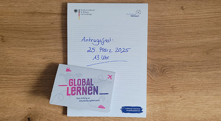 Notizblock mit handschriftlicher Notiz. "Antragsfrist 25. März 2025, 13 Uhr. Im unteren Drittel liegt ein Block mit Haftnotizen, auf dem Umschlag steht "Global Lernen. Von Anfang an. #AUsbildung weltweit".