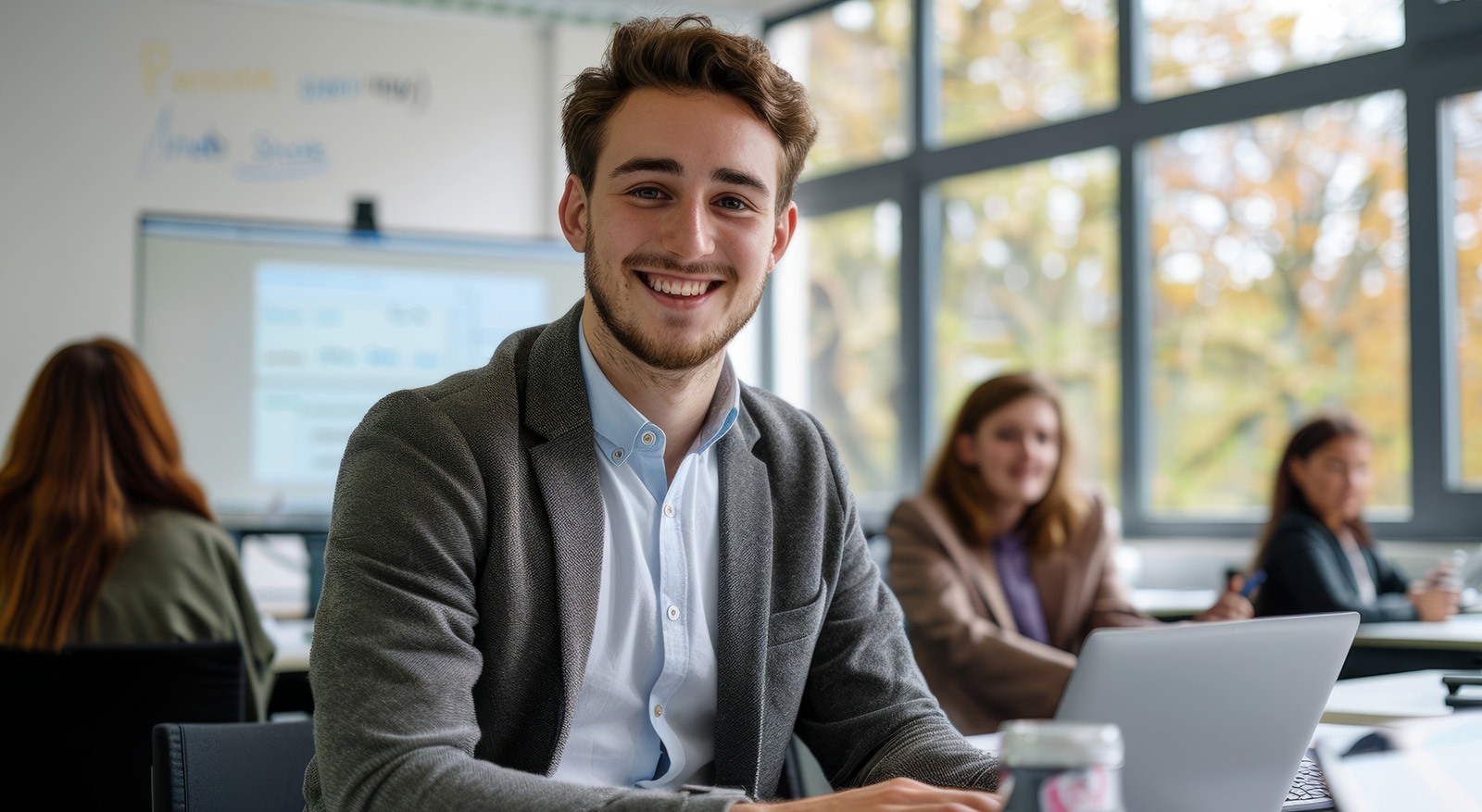 Lächelnder junger  Mann in einem Klassenzimmer, der an seinem Laptop arbeitet